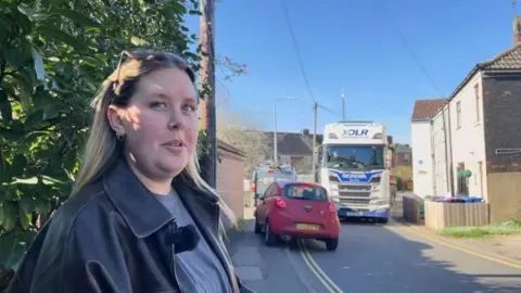 A woman looks concerned as she stands on a footpath by the side of a narrow residential road as a red car mounts the pavement to avoid white HGV lorry heading in the other direction. The woman has dark-blond hair and wears a black leather jacket, grey-blue T-shirt and sunglasses on the top of her head. Green shrubs and white-washed houses can be seen. The sky is blue.