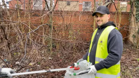 Dan Bater/BBC A man wearing a black hooded top with a yellow high-vis jacket. He is holding open a sack with someone using a litter picker to put rubbish in. In the background there are trees and leaves on the ground. 