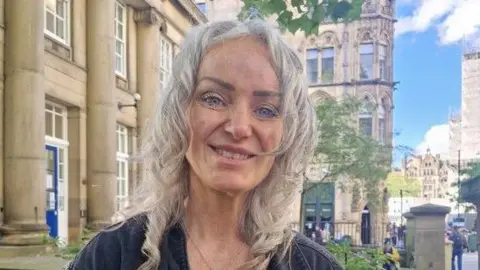 Gill Charlton, who is wearing a leopard print top and black denim jacket with tassels is smiling the camera with her hands grasped together. 