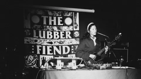Mark Buchanan A black and white photo of a woman with long, dark hair performing in front of a banner which reads The Lubber Fiend. The performer is singing into a microphone and playing an electric guitar. There is a table in front of her with various pedals and a laptop.