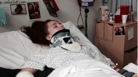 Harriet Barnsley A photo of a woman in a hospital bed wearing a cast around her neck. She is asleep in the photo and she is wearing a white hospital gown, and there are photos of her friends and family behind. 