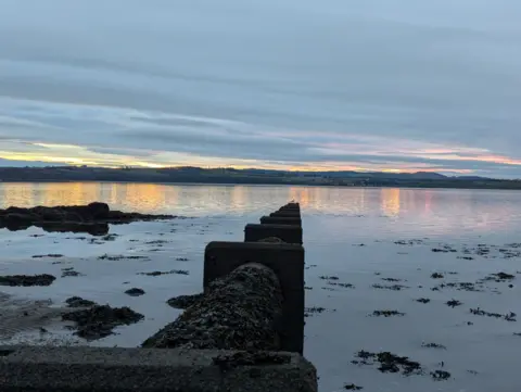 Alison Stewart Cloudy grey sky with pink and orange sunset visible in the far distance, above calm waters with the sunset reflecting on it next to a sandy beach covered in seaweed with a large pipe running through the centre of the image into the water.