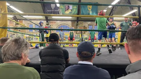 A group of primary school age children spar with each other in a boxing ring with green and gold ropes around it. Some men are sat in the club watching them from the sidelines. There are lots of pictures on the surrounding yellow walls.