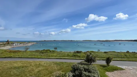BBC The image shows Grand Havre bay. There is a road and behind it is a stretch of green grass and then blue sea with moored boats. The sky is blue with white clouds. 