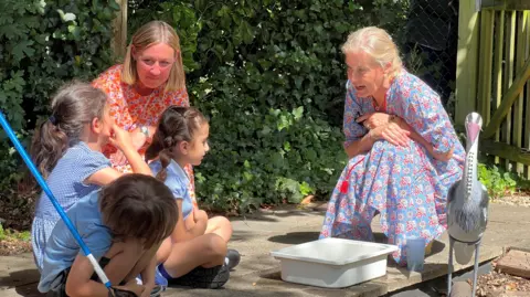 The Duchess of Edinburgh kneels down to talk to three young schoolchildren with a teacher watching on. Between them is a white box on the ground and a model of a heron.