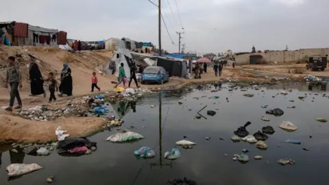 EPA People walk next to a sewage spill and garbage near tents for internally displaced people at a temporary camp in Rafah camp, southern Gaza Strip