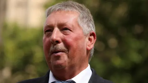 Getty Images Sammy Wilson speaking. He has grey hair and a moustache. He is wearing a white shirt and black jacket. The photo is cropped so you can only see his face and collar.