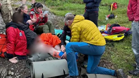 Keswick Mountain Rescue Team providing first aid to the injured volunteer. The volunteer is sitting on stony ground with his leg stretched out. A rescuer, wearing a yellow coat, is providing first aid. A number of other mountain rescue team members are standing or sitting nearby. Roll mats and hiking bags are lying on the ground.