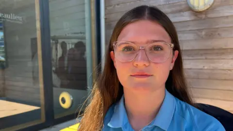 A girl with brown hair and clear glasses wearing a blue polo school uniform looking at the camera. Behind is a wooden building with a window to the left and a outdoor light on the right.