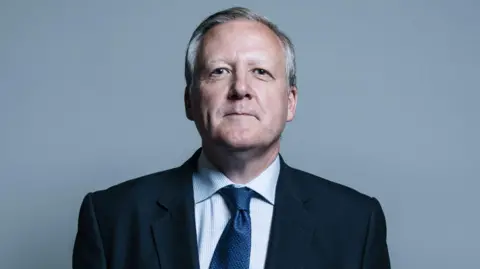 Kevan Jones, who wears a navy suit, tie and blue shirt. He sits and faces the camera with a grey background.