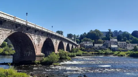 BBC Weather Watchers: Sunset Song A bridge over a river with some buildings on the far bank under a blue sky