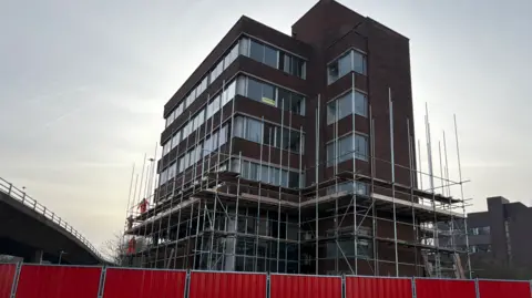 LDRS Computer House is a brown-brick multi-storey tower block near the Gateshead Flyover. Half of the building is covered in scaffolding. The structure is surrounded by a red a fence.