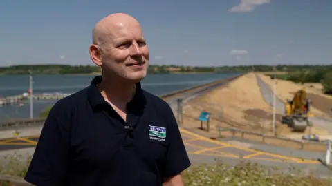 Doug Clarke, from Severn Trent, smiles as he looks to the side wearing a dark blue Severn Trent top. In the background Draycote Water reservoir can be seen.