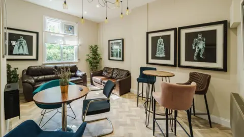 The Dynamo Project A room with two brown sofas against the walls, then a table with teal chairs and a higher table with three bar stools. The room has pale herringbone flooring and cream walls.