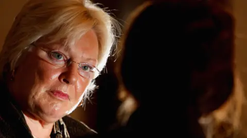 Getty Images A warmly lit indoor scene showing two individuals facing each other. Margo MacDonald with light-colored hair is in the foreground, partially turned toward another person who is seen in silhouette on the right. The background is softly lit with warm tones, creating a quiet, intimate atmosphere.