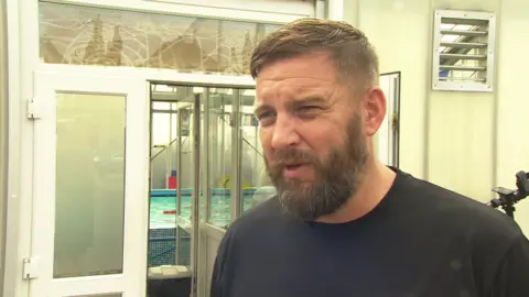 A white man stands outside a swimming pool looking off to the side of the camera talking.
