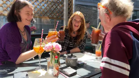 Three people sitting around an outdoor table, raising drinks in a toast. The table holds a flower vase with pink flowers, a tabasco sauce bottle, and some menus or papers. A lattice fence and part of a building are visible in the background.

