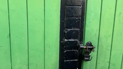 A wooden door, painted lime green, peppered with small holes which are said to be strike marks from shotgun pellets