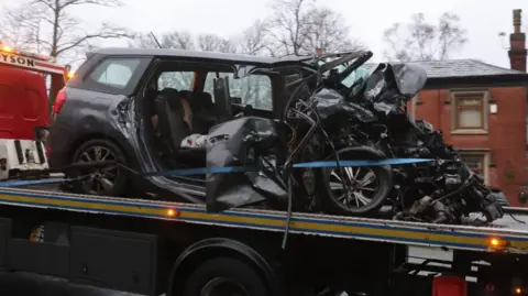 Phil Taylor A dark Seat car with its front bonnet completely smashed and both right doors torn off is towed away on the back of a lorry past houses.