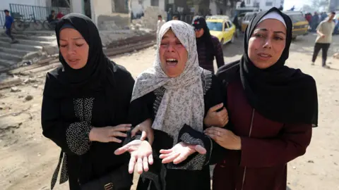 Three women linking arms with expressions of despair, with woman in the centre crying. Woman on left of image is wearing a black abaya with a black hijab, woman in centre, the same with a white printed hijab and the woman on the right is wearing a burgundy abaya with a black hijab and white bandana underneath it.