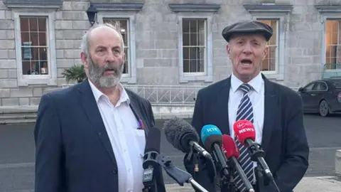 PA Media Brothers Danny and Michael Healy-Rae standing in front of a row of microphones. Danny is on the left - he has grey hair and a beard. He's wearing a white shirt and a blazer. Michael is wearing a suit, with a striped tie, and is wearing a flat cap