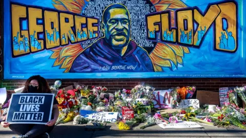 Getty Images Flowers, signs and balloons are left near a makeshift memorial to George Floyd near the spot where he died while in custody of the Minneapolis police, 29 May 2020