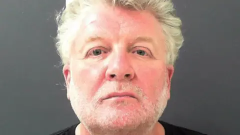 Close up shop of a man with a short white beard and white hair. He has grey eyes and is looking directly into the lens with a neutral, slightly sad expression.