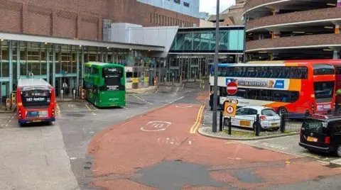 Eldon Square bus station. Two buses are parked in pays outside the glass doors of the entrance to the shopping centre, with two further buses parked behind them next to a circular multi-storey car park, with a white taxi next to them and a black cab behind. 
