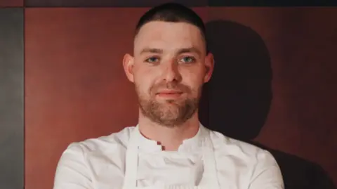 A man wearing a chef's white shirt and apron standing in front of a brown background.