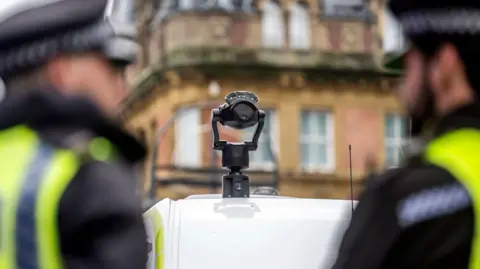 A black camera is mounted to the top of a white van. Two police officers are blurred in the foreground and there is an ornate building with windows in the background.