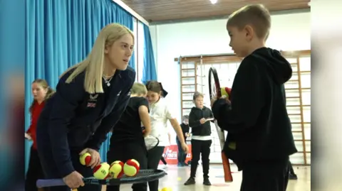 BBC The image shows Sonay Kartal talking to a child: she has a tennis racket in her right hand, which has 6 tennis balls on it, as well as one in her left hand. The boy she is speaking to is blonde and also holding a tennis racket and ball.