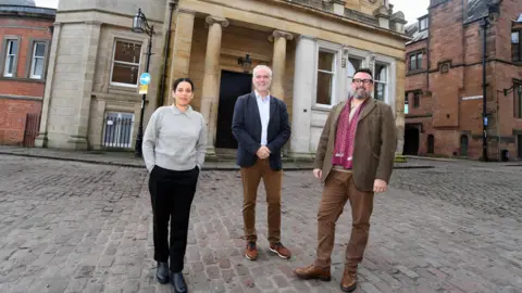 Richard Nelmes Three people stand in front of Drapers' Hall in Coventry. There is a woman with dark hair tied back, wearing a grey pullover, black trousers and black shoes. There is a man with short white hair wearing a dark blue jacket, a light blue shirt, brown trousers and brown shoes. A second man with short dark hair, glasses and a grey beard is wearing a brown jacket, brown trousers and brown shoes with a burgundy scarf.