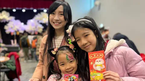 A woman and her two daughters pose for a photo, holding red packets.

