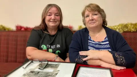 Two middle-aged women sit on a club sofa opposite each other with framed photographs and documents on the table. Julie, on the left, has long brown hair and wears a green Christmas T-shirt. Janet, on the right, has short, light brown hair and wears a blue and white patterned top and a dark blue cardigan. 