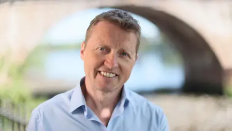 Mike Robinson smiling at the camera. He has short brown hair and is wearing a light blue open-necked shirt. He is standing next to a river and a bridge can be seen in the background.