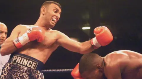 Prince Naseem Hamed punches with his opponent, who bows down to try to avoid the hit, with his left hand in a red boxing glove in the ring. He wears shorts in leopard print and the word Prince on his belt around the waist.