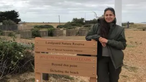 Contributed Ms Washington leans against a wooden information board at Landguard Nature Reserve. Her hair is partly tied back and partly down and she wears a green coat with a scarf and black trousers.