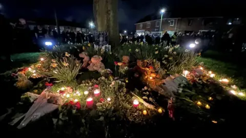 Dickon Hooper/BBC People gather around flowers left in tribute to three children who died in Sea Mills, Bristol