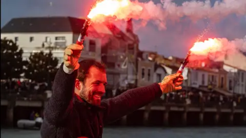 Pierre Bouras A man holding red flares in each hand smiles with a crowd in the background.