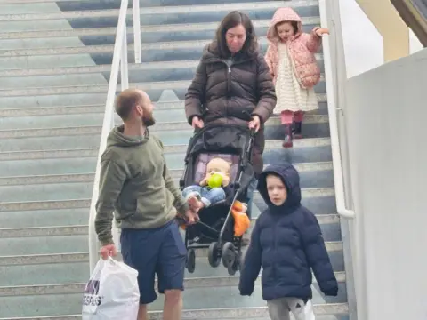 A man and woman carrying a child in a pram down steps, with two other children with them.