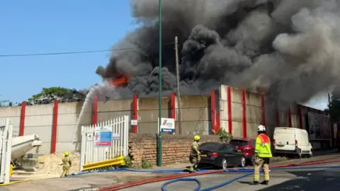 Nottinghamshire Fire and Rescue Service A large fire, with black smoke and flames coming from behind a concrete wall and firefighters squirting water