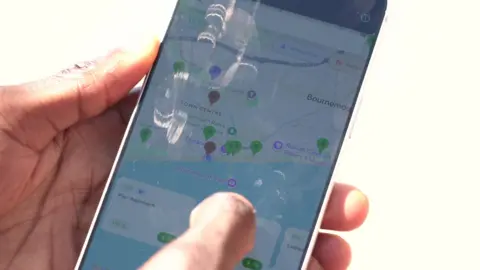 A smartphone being held in a man's hand, with the 'Where to Wee' app visible on the screen. Locations are highlighted with a coloured pin, either red, green or purple, marking where the public toilets are