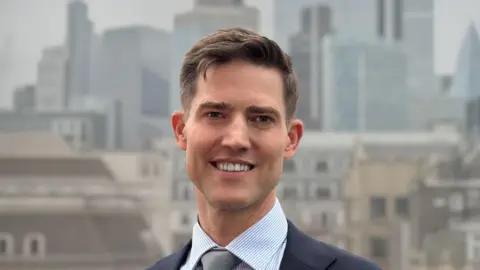 City of London Corportation A man with brown hair and wearing a navy blue suit jacket, a light blue shirt and a grey tie stands looking at the camera with the city of London in the background.