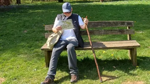 Roy Howard A man holding a map and a walking stick. He is wearing tracksuit bottoms, walking boots, a white t-shirt and a blue cap. 