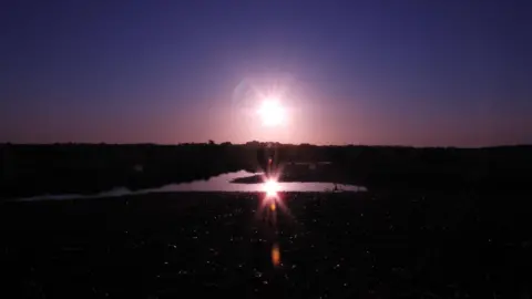 The sun is reflected in a pool of water. The sky is a beautiful purple colour while the rest of the picture is in darkness.