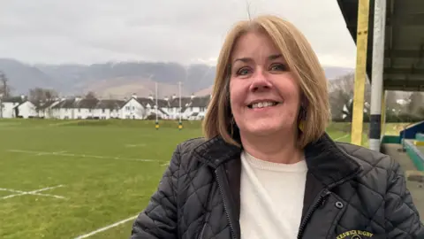 BBC/Ed Hanson Liz Weightman smiling into the camera. She has light brown, shoulder-length hair and is wearing a black padded coat and white jumper underneath. She is standing beside a rugby pitch with rows of white houses behind it. In the background there are mountains and the tops are covered in clouds.