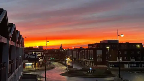 KELLY MCCORMACK/BBC A bright orange sunrise fills the sky above buildings and a roundabout in a darkened Ipswich town centre.