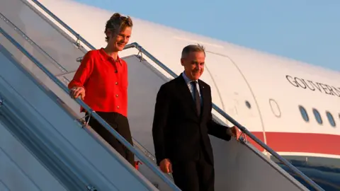 Canada's Prime Minister Mark Carney (right) and his wife Diana Fox Carney disembark an aircraft that says 'Gouvernement Du Canada' along with a red maple leaf, at Paris Charles de Gaulle airport on Monday morning against a clear blue sky. He wears a dark suit and tie and she wears a red shirt and black dress.