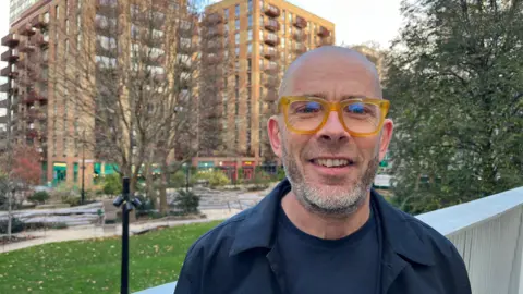 A man in glasses stands in front of a block of flats 