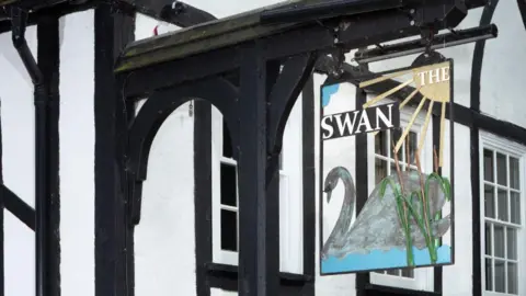 Historic England The white painted exterior of The Swan with black painted beams. A sign of a swan with writing which says THE SWAN in white lettering, hangs from the outside of the building.